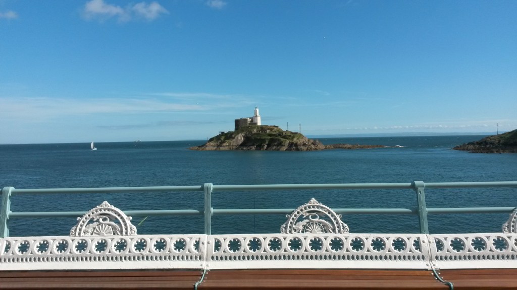 Photo showing an attractive painted white railing in the foreground, the sea and an island with a white lighthouse. The sky is blue above