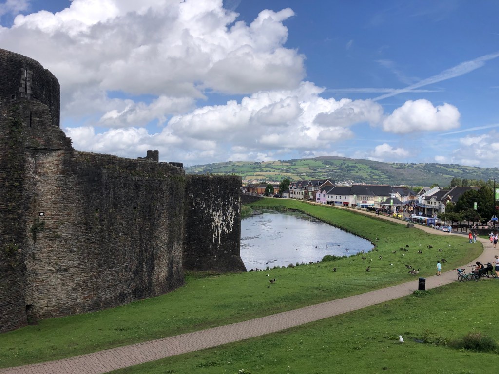 Photo of Caerphilly Castle in Wales. Images includes part of the castle on the left hand side, a moat filled with water and a green area with a path used by people. Houses followed by Welsh hills are in the background