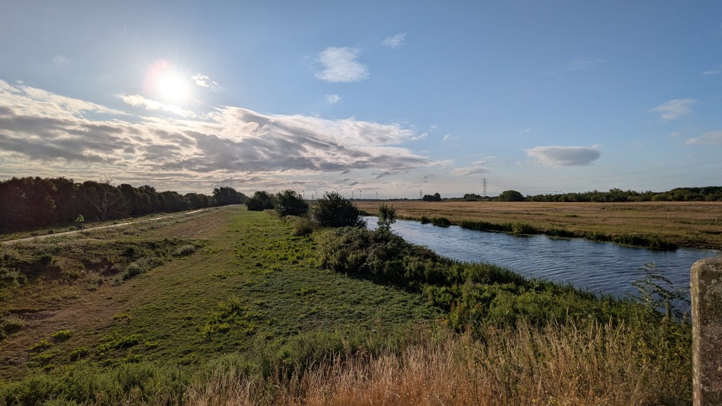 A photo of a flat landscape with a river, fields and a cycle path with a cyclist riding, under blue sky, clouds and sunshine.